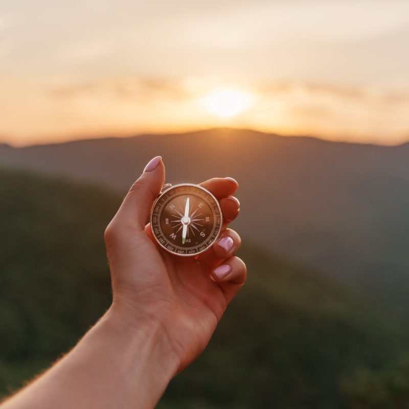 Explorer young woman holding compass in hand in summer mountains at sunrise, point of view. Concept of hiking and travel.